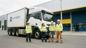 Quatre élèves en tenue de sécurité se tiennent devant un camion poids lourd et un entrepôt moderne d’un centre de formation transport-logistique à Arques.